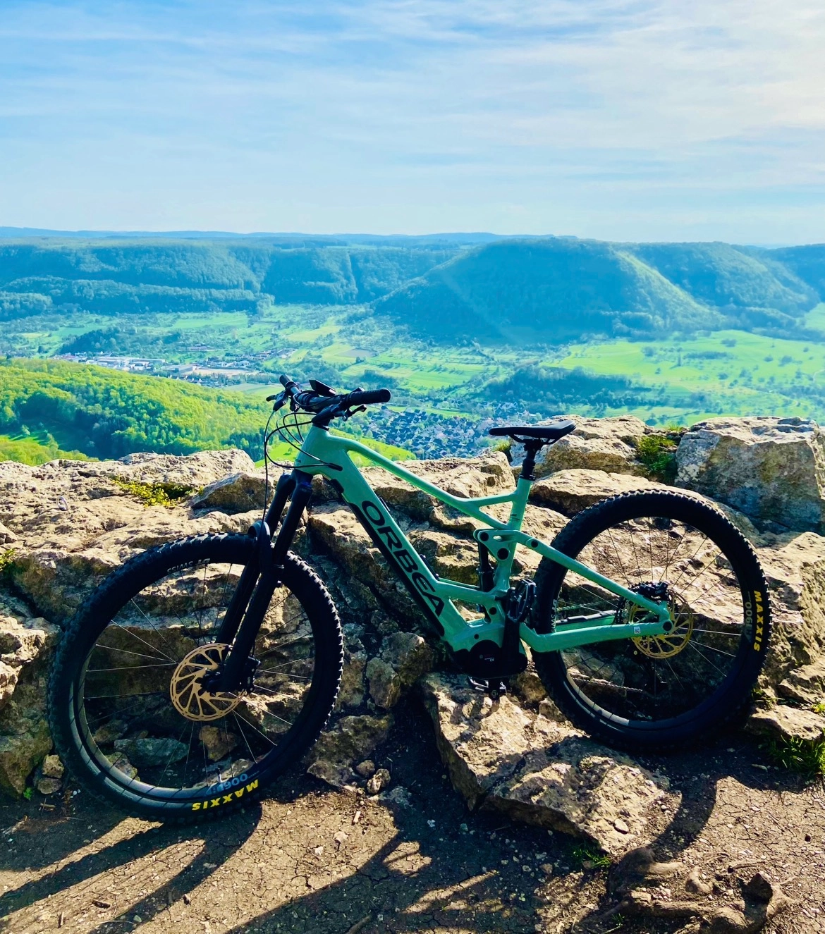 Grünes Mountainbike liegt auf einen Felsen vor einer tollen Aussicht auf grüne Bäume in einer Schlucht mit blauem Himmel und Sonne.
