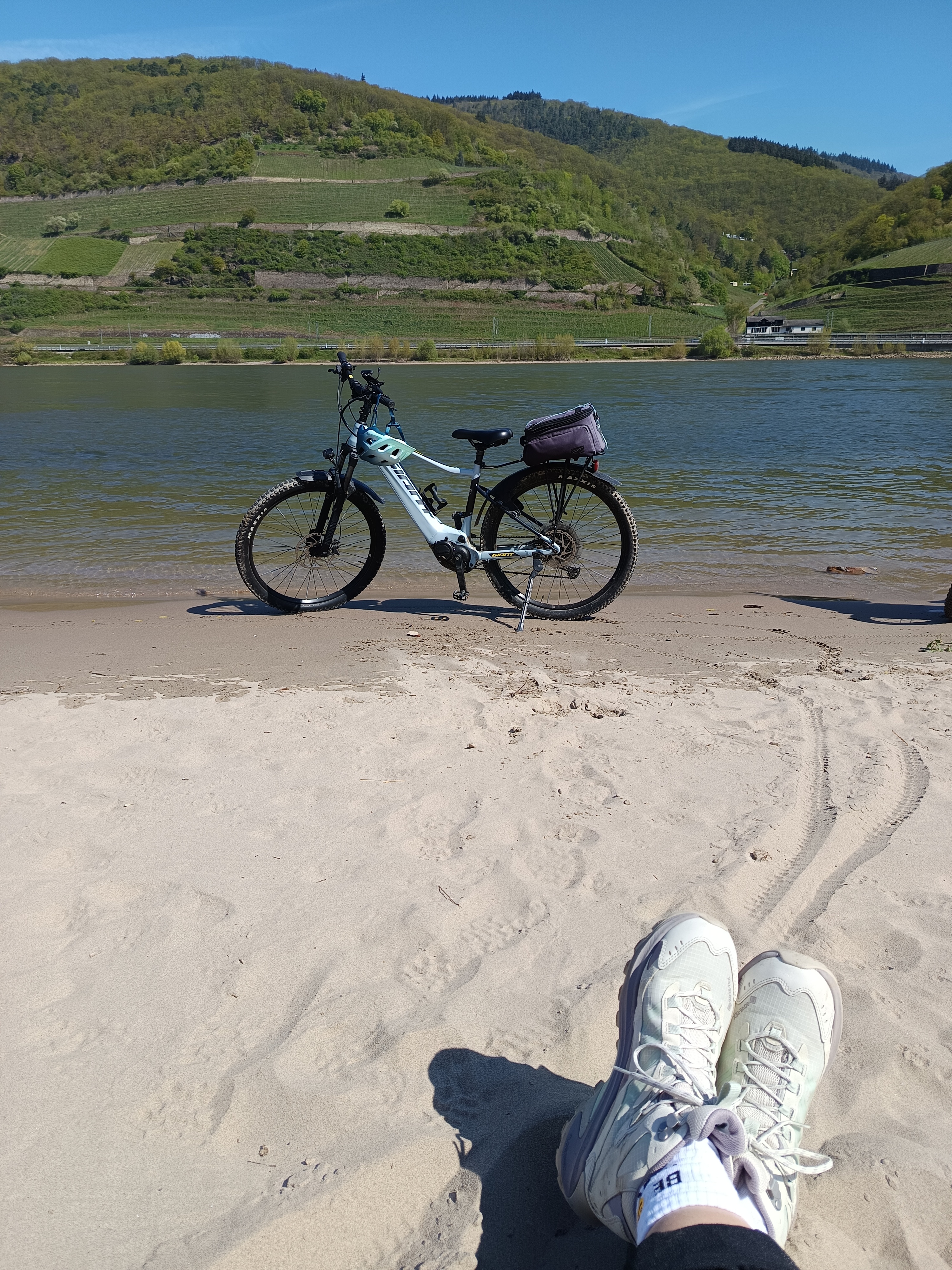 Person sitzt am sandigen Flussufer, Beine mit Sportschuhen im Vordergrund; daneben steht ein Fahrrad, gegenüber liegen grüne Hügel und Weinberge.